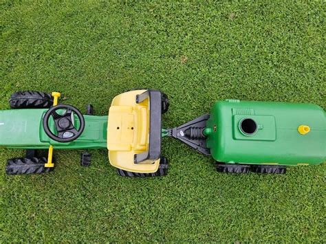 Fragment traktora Rolly Toys John Deere pokazujący otwartą maskę silnika oraz ruchomą łyżkę koparki.