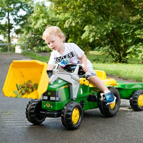 Photo of a green John Deere RollyKid pedal tractor with a front loader and an attached trailer, on a grassy background.
