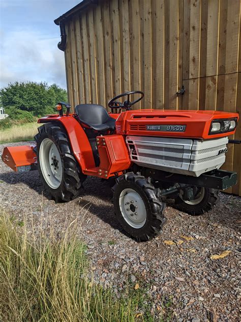 The Kubota B1600 compact tractor is shown working in a garden, pulling a small trailer. The image highlights its compact size and versatility for various tasks.