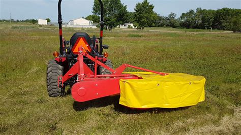 Photo of a tractor-mounted rotary drum mower in action, cutting tall grass in a field