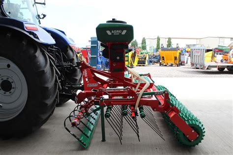 The AGRO-MASZ Grass 300 seed drill in action on a field, showing its components like harrows and rollers.