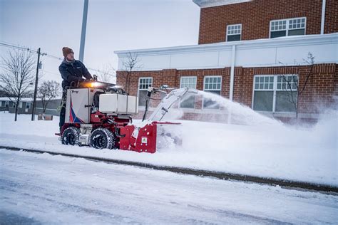 Thematic photo of a person operating a 100cm wide gas-powered sweeper/snow blower on a large paved area, removing leaves or light snow.