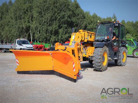 Pług kątowy JCB zamontowany na koparko-ładowarce, z zaznaczonym mechanizmem uchylnym i płozami ślizgowymi.